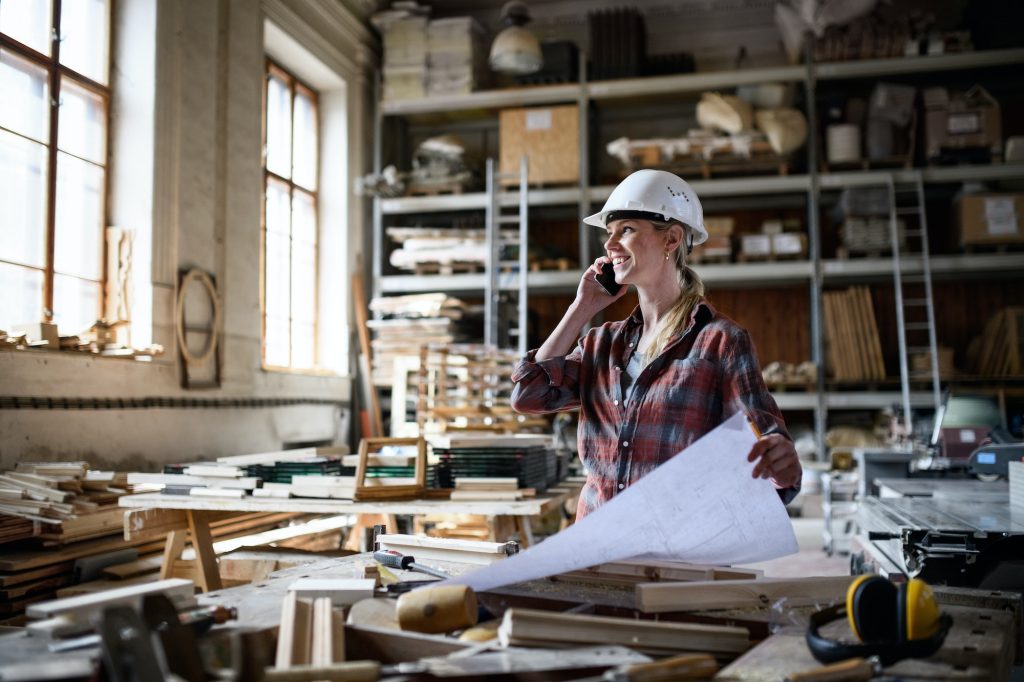 Female engineer holding blueprints and making phone call indoors in carpentry workshop