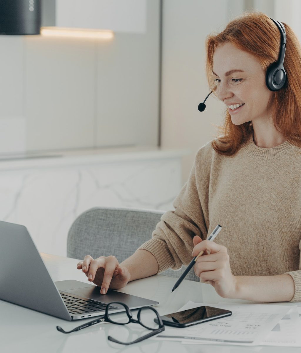 Happy redhead woman watches online webinar uses headset focused at laptop screen